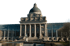 Bayerische Staatskanzlei, ein großes, klassisches Gebäude mit Kuppel, Säulen und Glasflügeln, vor grünem Hintergrund.