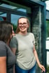 Eine lächelnde Frau mit Brille und T-Shirt unterhält sich mit einer anderen Person vor einem modernen Gebäude.