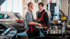 Zwei Mechaniker in Overalls unterhalten sich in einer Autowerkstatt, ein Auto steht links.