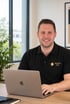 Lächelnder Mann im schwarzen Mission Kita Poloshirt arbeitet am Laptop in einem hellen Büro.