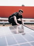 Ein Mann hockt auf einem Dach und greift nach einem Solarmodul, mit einem roten Gebäude im Hintergrund.