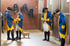 Vier Reiter in blau-gelben historischen Uniformen und Dreispitzhüten bereiten sich in einem Pferdestall vor.