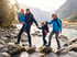 A family of four wearing hiking gear crosses a rocky river in a mountain valley.