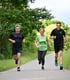 Drei Läufer, zwei Männer und eine Frau, joggen lächelnd auf einem asphaltierten Weg durch grüne Natur.