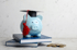 Blaues Sparschwein mit Doktorhut auf zwei Büchern, daneben ein Stapel Münzen.