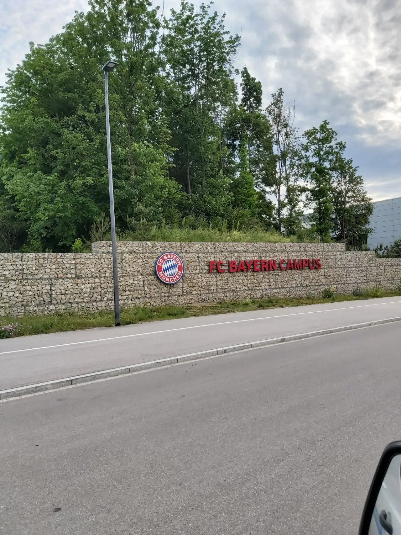 FC Bayern Campus Schild an einer Steinmauer mit Bäumen im Hintergrund und einer Straße davor.