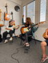 Two girls play electric guitars in a room with guitars on the wall and a window on the right.