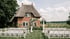 Hochzeit im Freien mit weißen Stühlen vor einem Backsteinhaus mit Strohdach und Blumenbogen.
