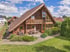 Modernes Holzhaus mit großer Glasfront, Balkon und Terrasse in einer Gartenanlage unter blauem Himmel.