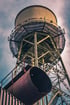 Ein industrieller Wasserturm in Bochum auf einer Stahlkonstruktion mit einem großen Rohr im Vordergrund vor bewölktem Himmel.