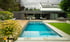 Moderner Garten mit rechteckigem Pool, Holzterrasse, Rasenfläche und Haus mit großen Glasfenstern unter bewölktem Himmel.