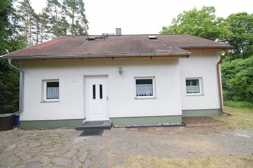 Weißes Einfamilienhaus mit rotem Dach, grüner Basis und weißer Tür, umgeben von Bäumen.