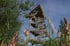 Hölzerner Aussichtsturm mit Wendeltreppe vor blauem Himmel, umgeben von grünen Bäumen und rosafarbenen Blumen.