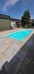 Rechteckiger Außenpool mit türkisblauem Wasser, umgeben von hellen Natursteinplatten und einem grünen Wohnhaus.