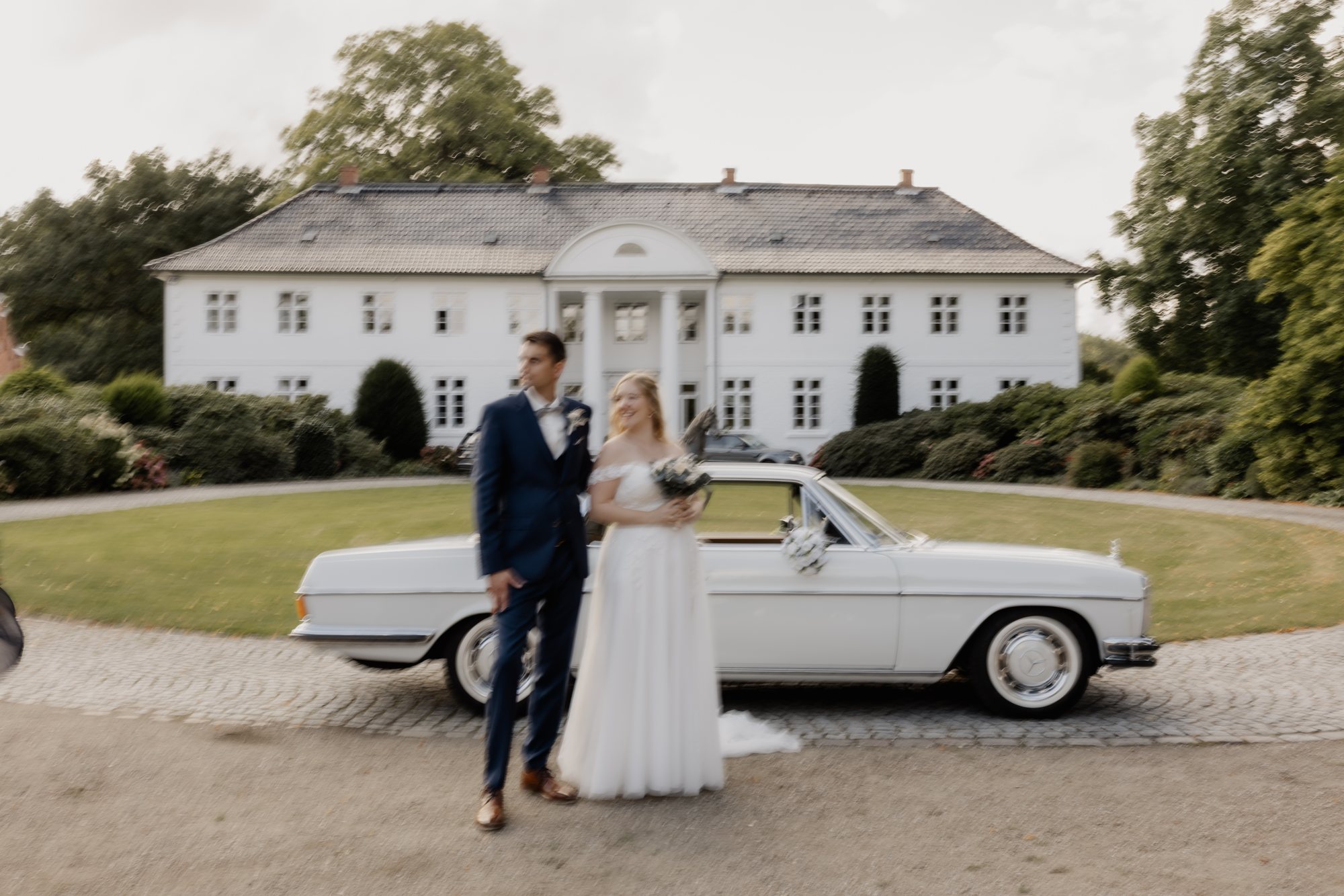 Brautpaar steht vor einem weißen Oldtimer in der Auffahrt eines Gutshauses