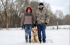 Paar mit Golden Retriever im Schnee, kahle Bäume im Hintergrund.
