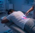 Man lying on stomach receiving laser therapy on his lower back from a therapist.