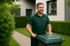 Mann in grüner Arbeitskleidung schiebt eine grüne Mülltonne auf einem Gartenweg.
