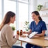 A smiling healthcare professional in blue scrubs shows data on a tablet to a patient during a medical consultation.