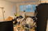 Unaufgeräumtes Zimmer mit Müll auf dem Boden, einem weißen Sofa, Standventilator und schwarzem Regal vor einem Fenster.