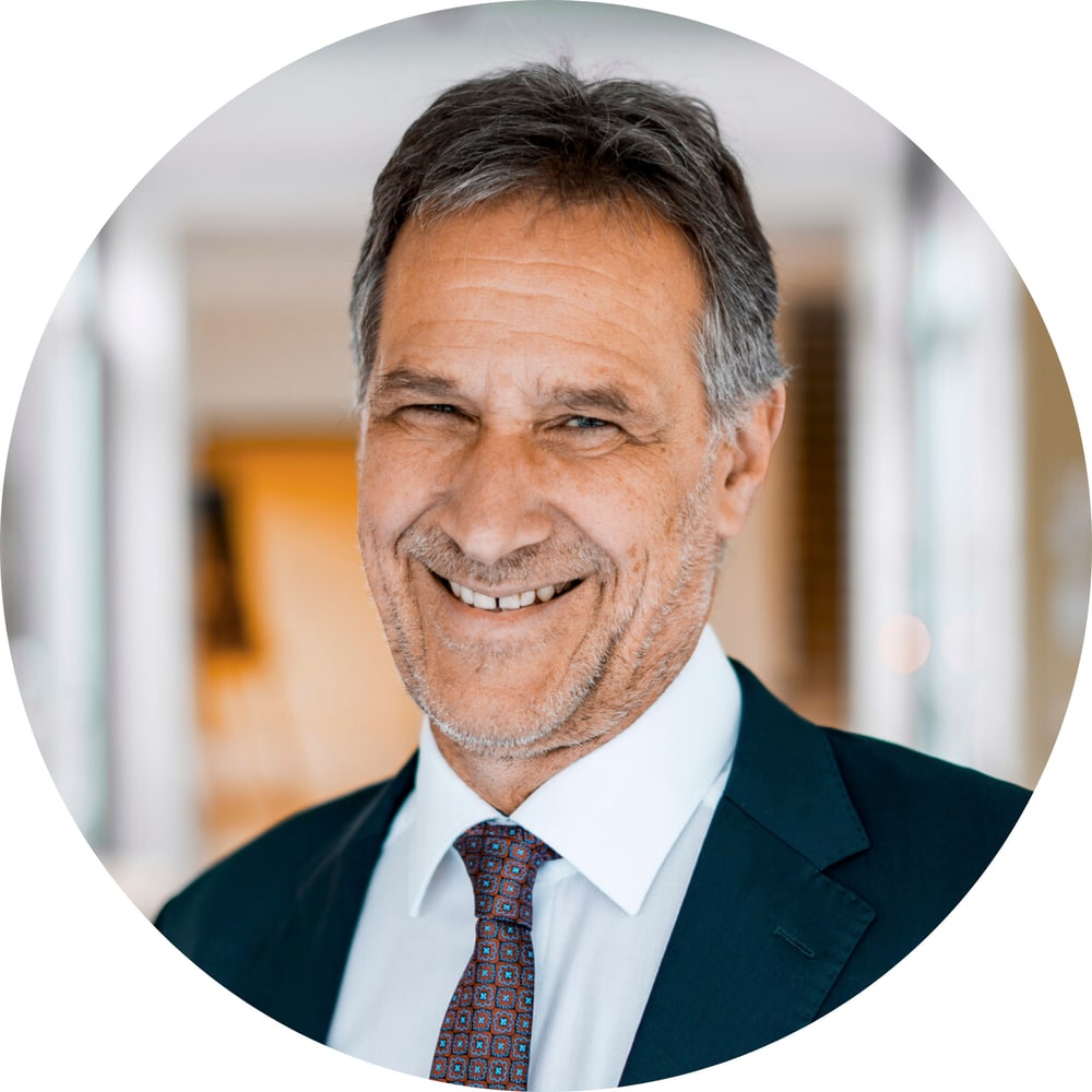 Ein lächelnder Mann mittleren Alters mit grauen Haaren, Anzug und Krawatte blickt nach vorne.