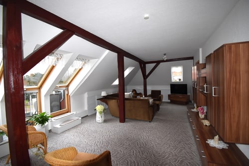 Geräumiges Dachgeschoss-Wohnzimmer mit dunklen Holzbalken, braunem Sofa, Sessel und Fenstern.