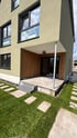 Modernes Einfamilienhaus mit grüner Fassade, überdachter Terrasse mit Holzverkleidung und Rasen mit Trittsteinen.