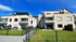 Zwei moderne Mehrfamilienhäuser mit Balkonen und Rasenflächen, getrennt durch einen Zaun unter blauem Himmel.