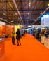 Menschen auf einer Messe mit orangefarbenem Teppich, Ständen mit "LA EVOLUCIÓN" und "Next Consulting".