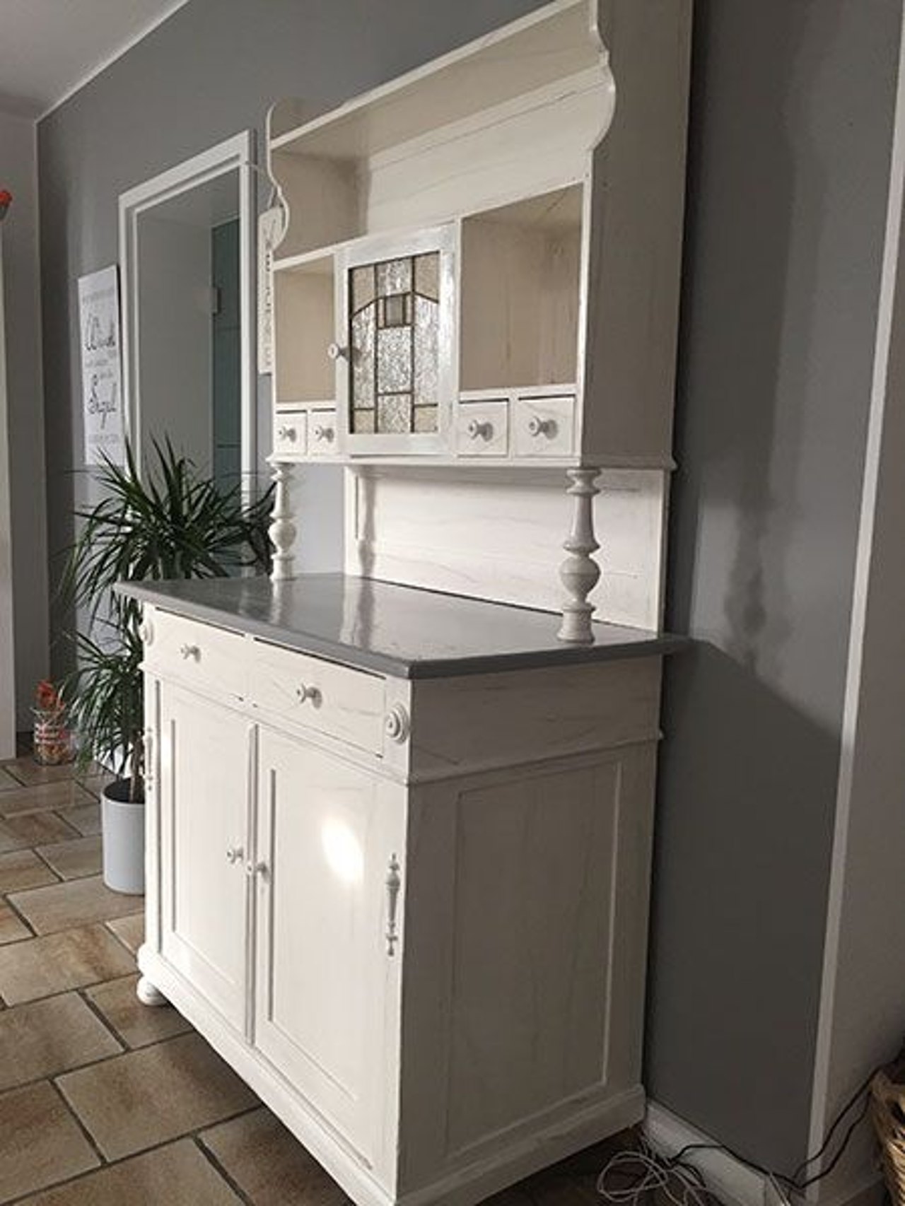 Weißes Buffet im Landhausstil mit grauem Oberteil, Bleiglasfenster und kleinen Schubladen vor einer grauen Wand.