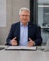 Ein lächelnder Mann mit Brille und Anzug sitzt an einem Schreibtisch mit offenen Ordnern und einem Laptop.