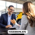 Mann im Anzug lächelt und spricht mit Frau am Tisch in einem Autohaus, gelbes Auto im Hintergrund. HAERBERLEN.