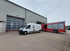 Weißer Transporter mit Firmenlogo und rotes Feuerwehrfahrzeug parken vor einer Industriehalle mit Rolltoren.