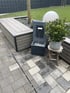 Moderner Gartenbrunnen aus grauem Stein neben einer Holzbank, einer Topfpflanze auf einem Hocker und einer Kugelleuchte.