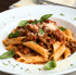 Penne mit Bolognese-Sauce, Parmesan und Basilikumblättern auf weißem Teller serviert.