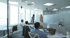 A business meeting in a modern office with five people, two standing by a whiteboard and three seated at desks.
