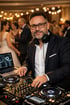 DJ mit Kopfhörern und Brille lächelt, während er an einem Mischpult mit Laptop und tanzenden Gästen im Hintergrund steht.