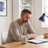 A bearded man in a tan sweater sits at a wooden desk looking down at an open planner next to a coffee mug and desk lamp.