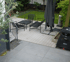 Eine Holzterrasse mit schwarzen Gartenmöbeln, Sonnenschirm und Grill; dahinter Rasen und Bäume.