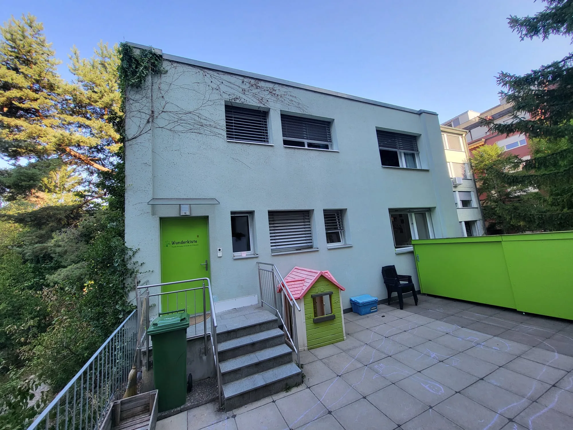 Das Kita-Gebäude mit grüner Eingangstür, Treppe und Terrasse.