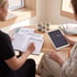 A nutritionist writes in a health guide journal while a client holds a tablet displaying data charts near herbal supplement jars.