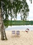 Zwei leere Liegestühle am Sandstrand, im Hintergrund ein See mit Wald und ein Rettungsring.