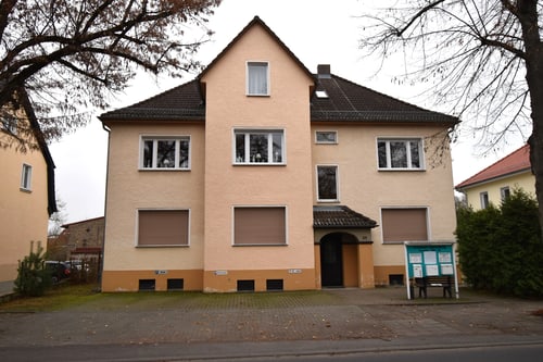 Ein dreistöckiges, beiges Wohnhaus mit braunen Fensterläden und einem schwarzen Eingangsbereich unter einem Vordach.