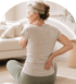 A woman sits cross-legged on a rug, holding her neck and lower back in a bright living room.