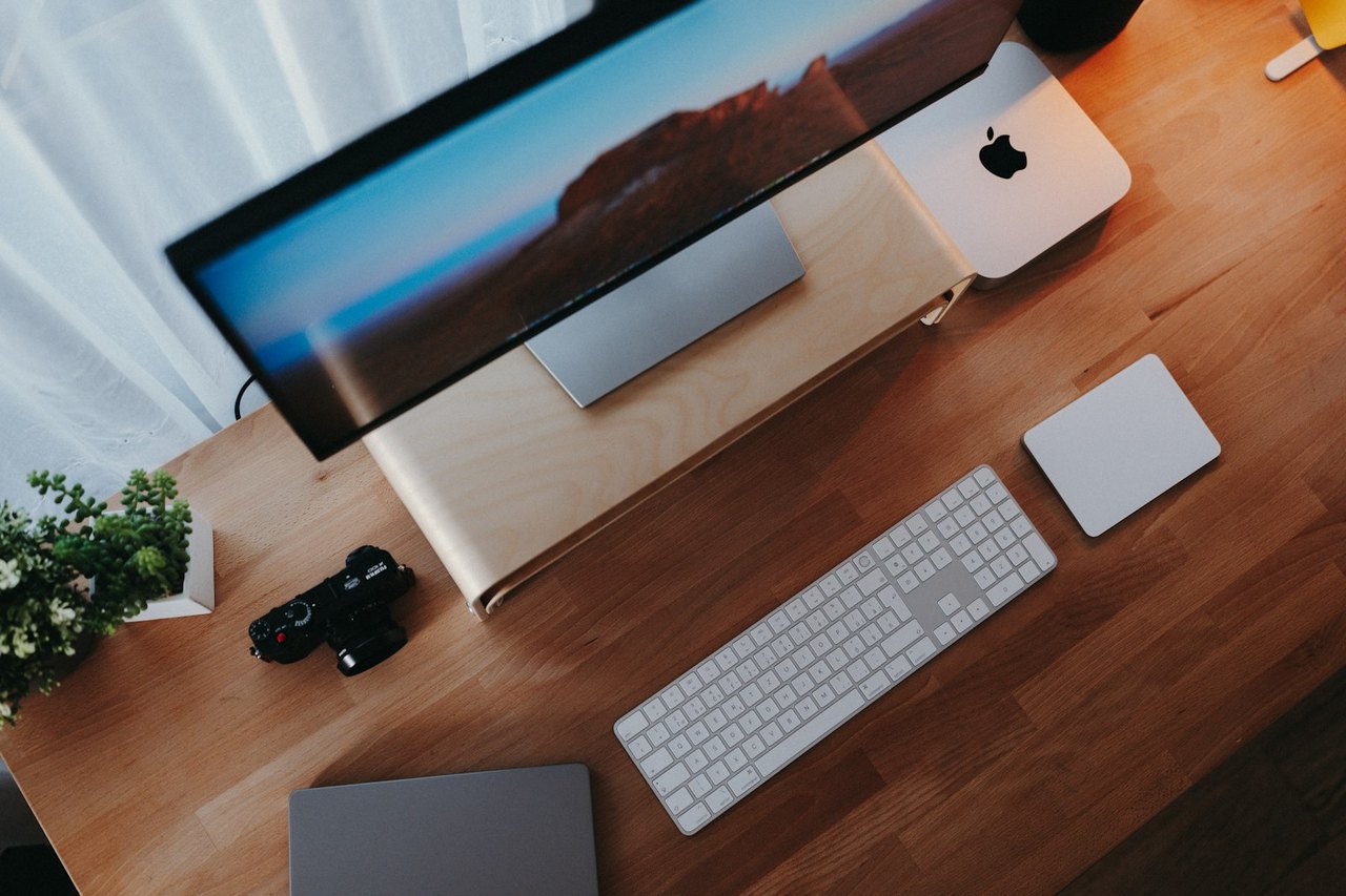 Schreibtisch von oben mit Bildschirm, Mac mini, weißer Tastatur und Trackpad auf Holzplatte