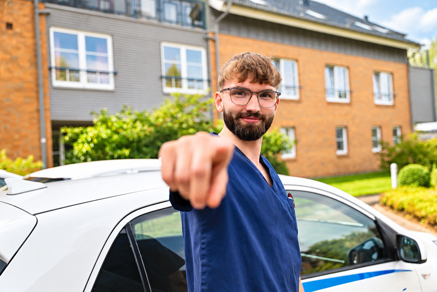 Ein Mann im blauen Kasack steht neben einem Fahrzeug und zeigt in die Kamera.