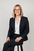 Frau mit blondem Bob, Brille und dunklem Anzug lächelt und sitzt auf einem Hocker.