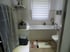 Helles Badezimmer mit weißer Badewanne unter einem Fenster mit Jalousie, einer Toilette und einem gemusterten Vorleger.