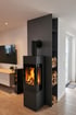 Moderner schwarzer Kaminofen mit brennendem Feuer vor einer dunklen Wand mit integriertem Holzregal im Wohnzimmer.