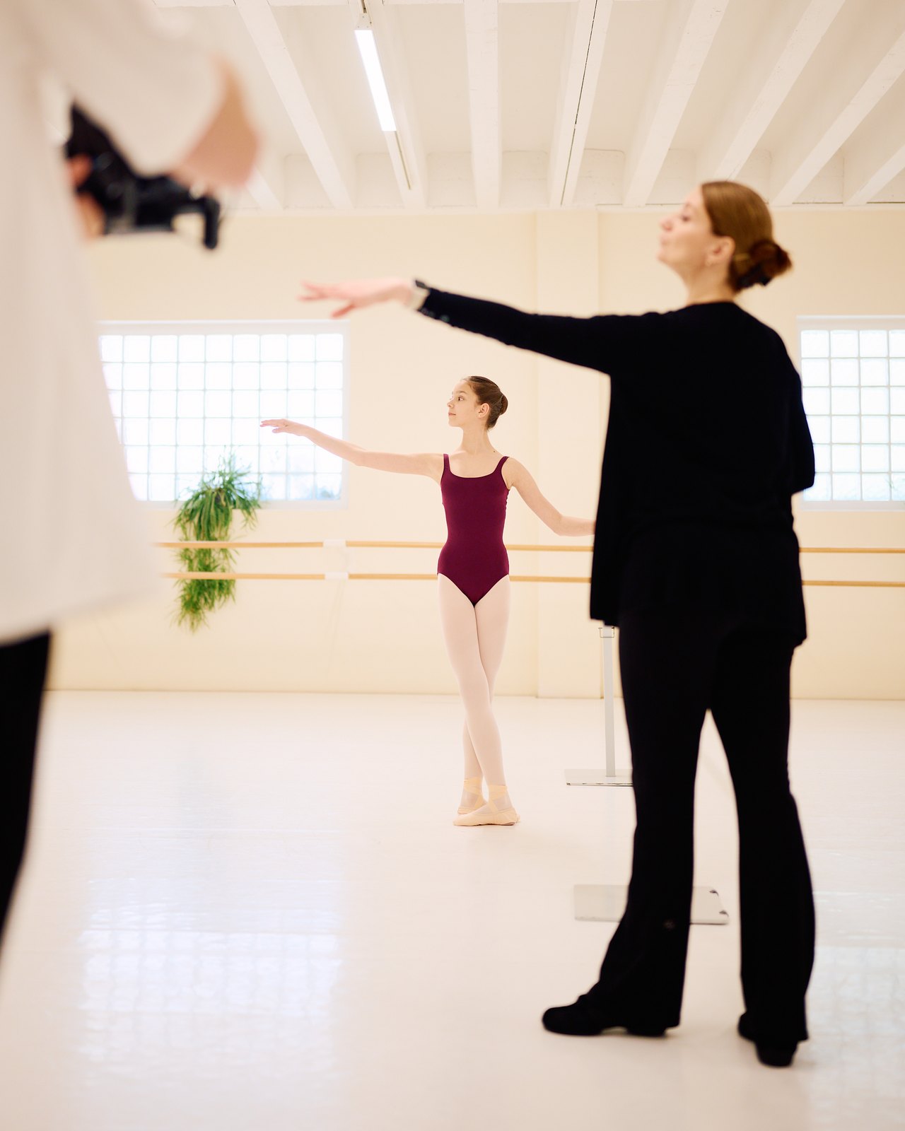 Ein Coach demonstriert in einem hellen Studio eine Armhaltung, während eine junge Tänzerin diese nachahmt; im Vordergrund ist eine Kamera zu sehen.
